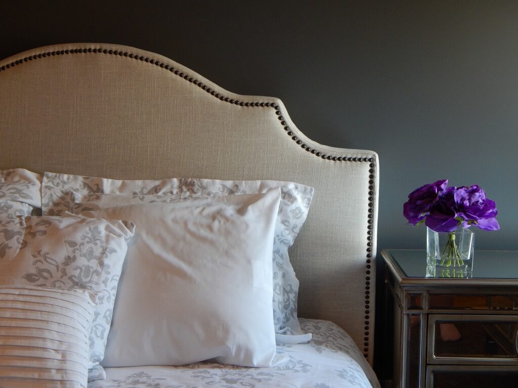 8 Simple DIY Home Projects For Beginners - Beige upholstered bed headboard with stud accents, white and floral-patterned pillows, and purple flowers in a glass vase on a mirrored nightstand.