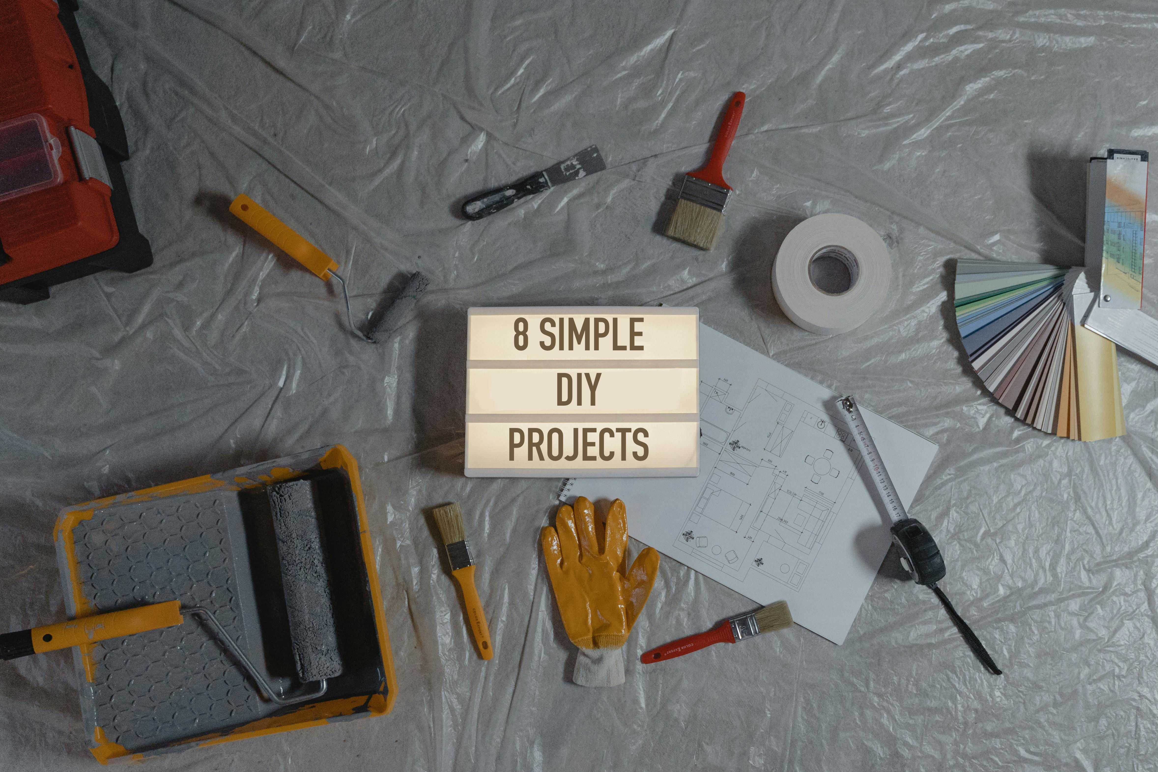 8 Simple DIY Home Projects For Beginners - Top view of essential tools for 8 simple DIY projects, including paintbrushes, roller, gloves, paint swatches, and blueprint layout on a plastic-covered surface